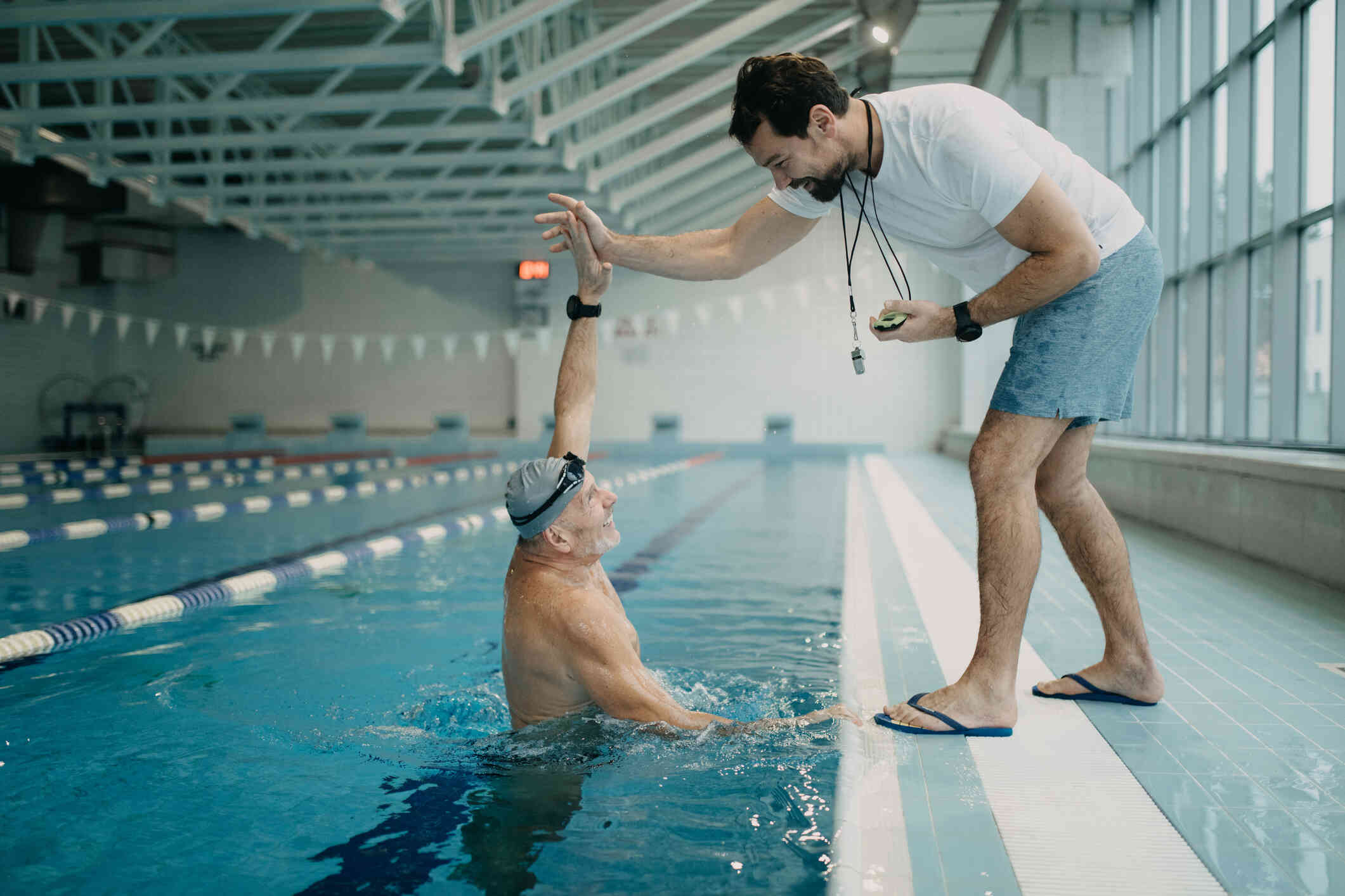 A man in a white shirt with a stopwatch highfives an elderly man in the pool with a smile.